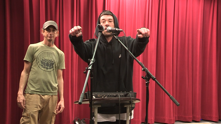 Red curtain background with a man dressed in a black hoodie in front of two mics. Man in a khaki tee and hat standing in background to the left.