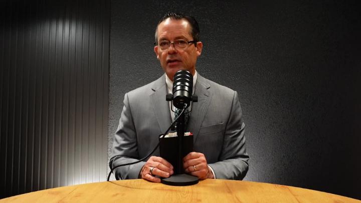 Man in a suit and glasses seated at a table behind a podcast mic. Dark walls behind