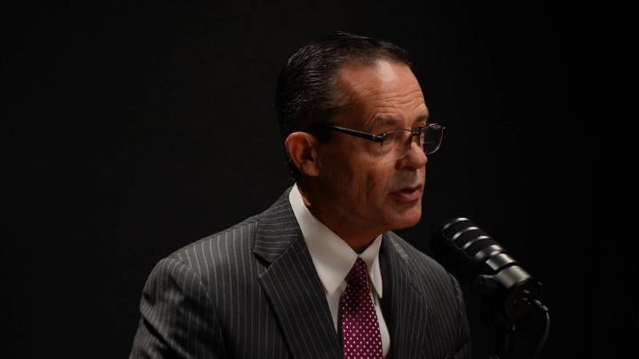 Man wearing glasses and a grey suit is in front of a podcast mic.