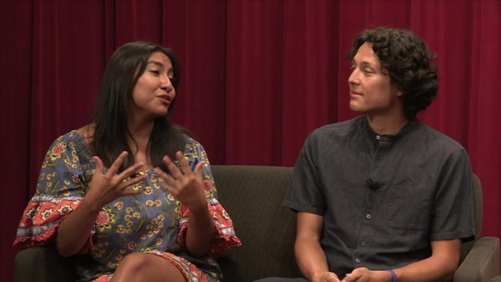 Red curtain background. Woman in a floral dress seated on the left. Man in a button up shirt seated on the right.