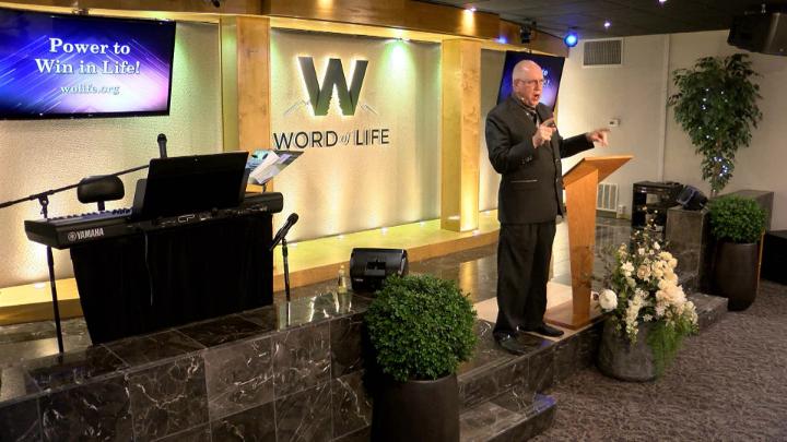 Man in a suit holding a bible next to a pulpit in the middle of a platform. Lit up white walls in the background with a large W and Word of Life in the center.