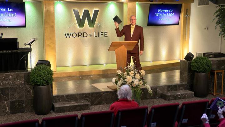 Man in a suit holding a bible next to a pulpit in the middle of a platform. Lit up white walls in the background with a large W and Word of Life in the center.