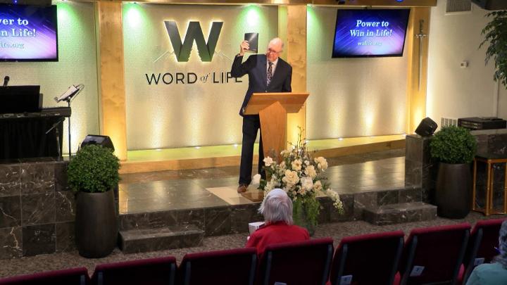 Man in a suit holding a bible next to a pulpit in the middle of a platform. Lit up white walls in the background with a large W and Word of Life in the center.