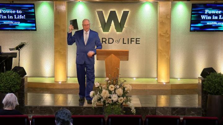 Man in a suit holding a bible next to a pulpit in the middle of a platform. Lit up white walls in the background with a large W and Word of Life in the center.