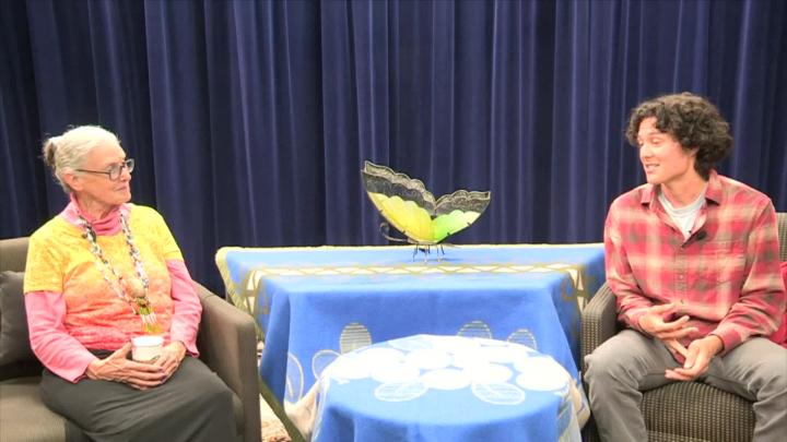 blue curtain background, blue tableclothed table in the center with a butterfly statue on it. Older woman in a yellow and orange top to the left. Man in a red plaid shirt seated to the right.