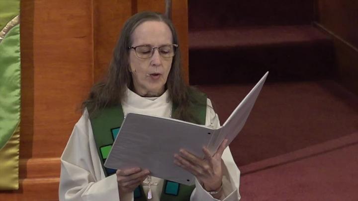Female priest in a white robe and green stole, holding a book.