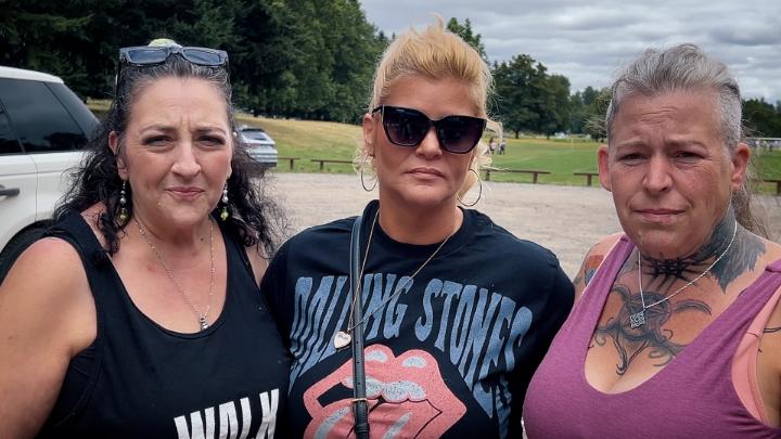 Outside picture. Woman on left in a black tank top. Woman in middle wearing sunglasses and a black tee. Woman on the right with tattoos and a pink tank top. 