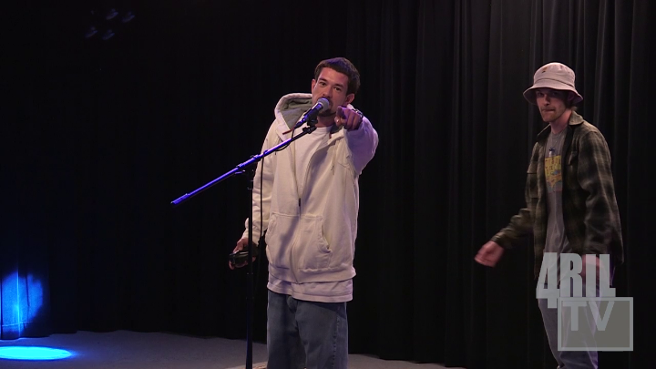 Black background. Man in a flannel and hat standing to the right. Man in the middle with a white shirt on, standing behind a mic pointing. Blue "4Ril" logo bottom left.