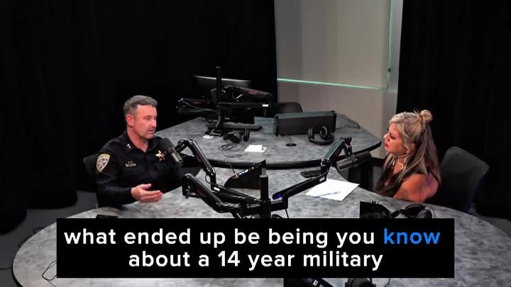 Man seated at a podcast table in a police uniform, to the left. Woman seated behind same table to the right.