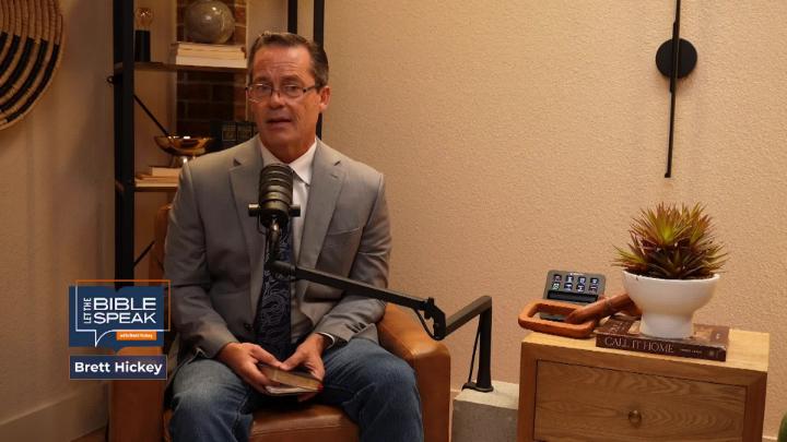 Yellow toned room. Bookshelf in the back left. Man in a grey suit seated behind a podcast mic.