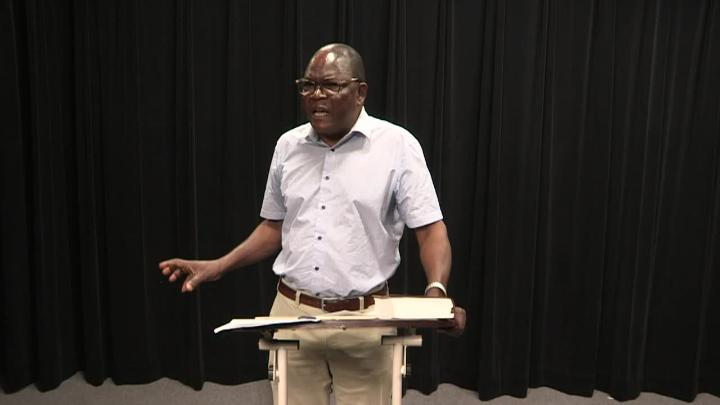 Black curtain background. Dark skinned man in a light shirt standing in the center behind a podium