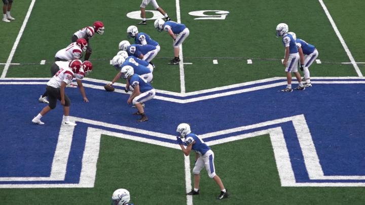 Football field with large blue "M" on it. Playes in blue and white uniforms and red, white and black uniforms lined up.