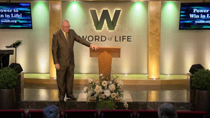 Man in a suit holding a bible next to a pulpit in the middle of a platform. Lit up white walls in the background with a large W and Word of Life in the center.