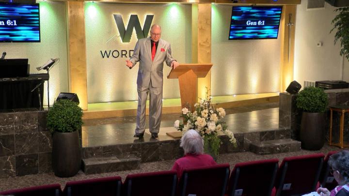 Man in a suit holding a bible next to a pulpit in the middle of a platform. Lit up white walls in the background with a large W and Word of Life in the center.