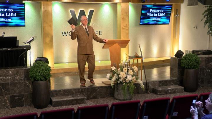 Man in a suit holding a bible next to a pulpit in the middle of a platform. Lit up white walls in the background with a large W and Word of Life in the center.