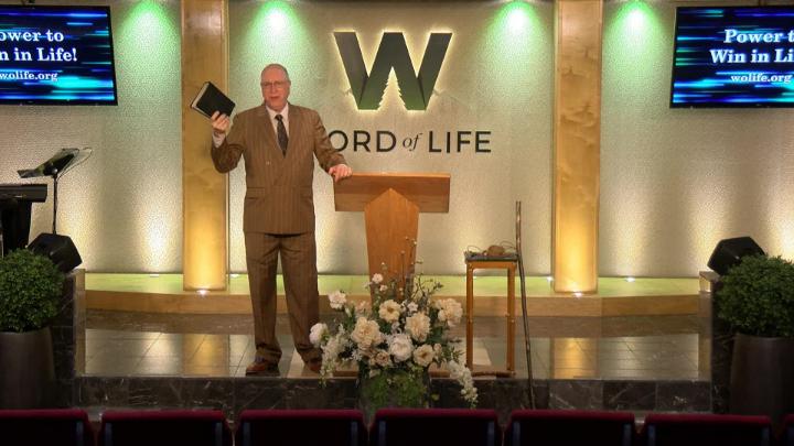 Man in a suit holding a bible next to a pulpit in the middle of a platform. Lit up white walls in the background with a large W and Word of Life in the center.