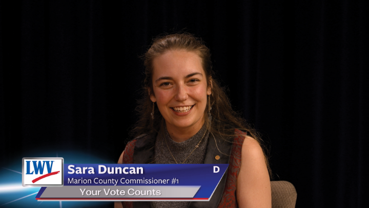 Black background. Woman in a sleeveless grey sweater with a red and black brocade vest in the middle. Lower thirds to the bottom left with red, white and blue LWV logo. White words on a blue background: Sara Duncan Marion County Commissioner #1 Your Vote Counts D
