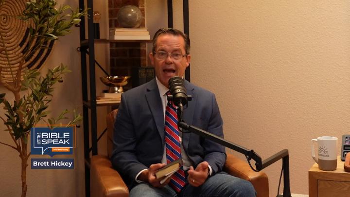 Yellow toned room with a knick knack shelf in the back left corner. A mand in a blue suit and tie is seated in front of a podcast mic just left of center.