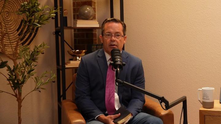 Man in a grey suit seated in front of a podcast mic. Yellow room with a knick knack shelf in the back.