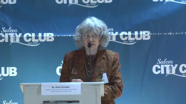 Blue background with white "Salem City Club" logo scattered on it. Older woman in a brown suit jacket standing behind a podium with a mic.