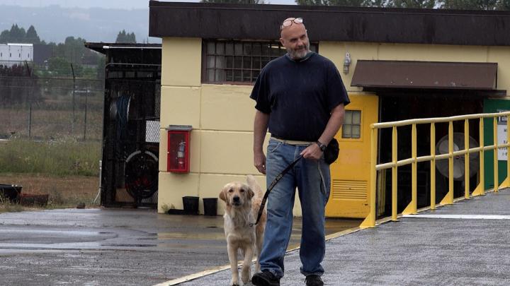 Outside picture of a man in the center wearing jeans and a black tee holding a leash to a lab looking dog on his left. A yellow building and fencing in the background.