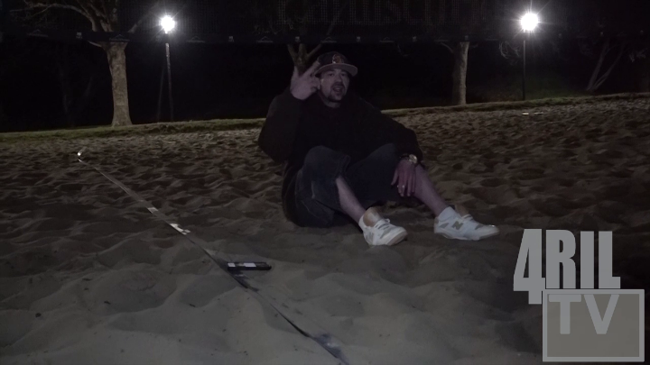 Man in black sitting on sand. White words lower right corner: "4Ril"