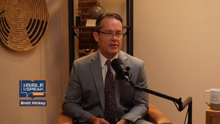 Man in a grey suit and glasses seated behind a podcast mic. Knick knack shelf in the background. Blue and orange "Let the Bible Speak" logo in lower left corner.
