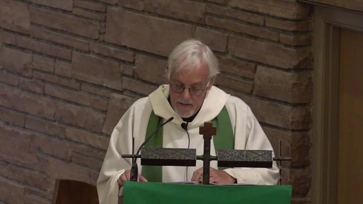Priest in white with a green stole standing behind a pulpit. Brick background.