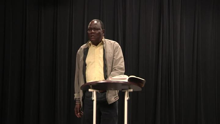 black curtain background with a dark skinned man in a yellow shirt and brown jacket standing in the middle behind a podium