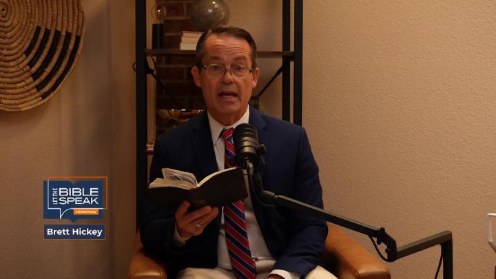 Yellow tone to the entire picture. Man in a suit seated in front of a podcast mic holding a bible. Knick knack shelf in the background. Blue and orange Let The Bible Speak logo bottom left.