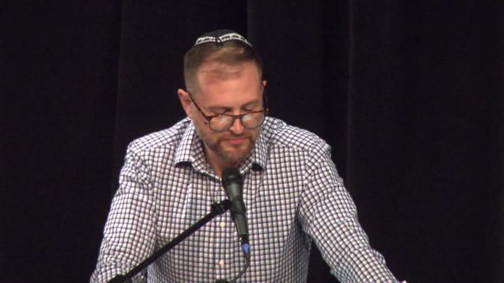 Black background. Man in a checkered shirt and glasses standing in front of a mic.