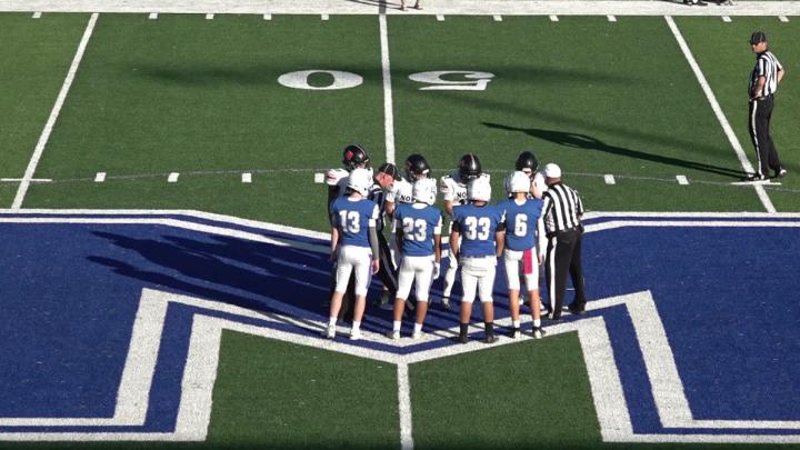 Football field with large blue "M" in the middle. Players wearing blue and white in the middle.