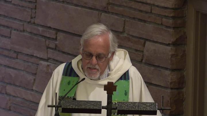 Priest with a white robe and green stole standing in front of a podium. Brick wall in the back.