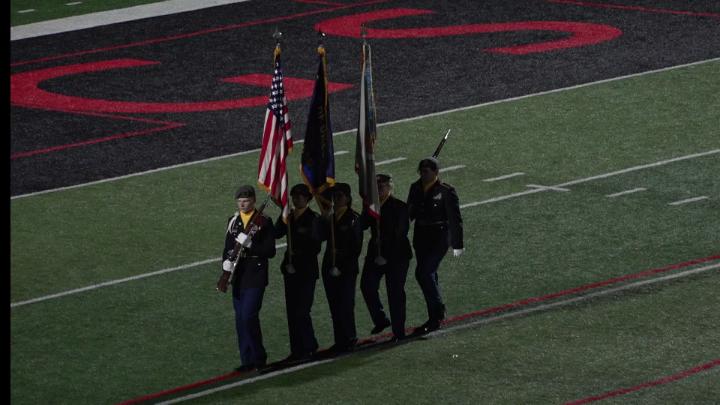 football field with color guard walking across holding the US flag