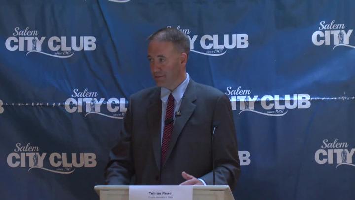 blue background with white Salem City Club logo on it. Man in a dark suit standing in the middle behind a podium.