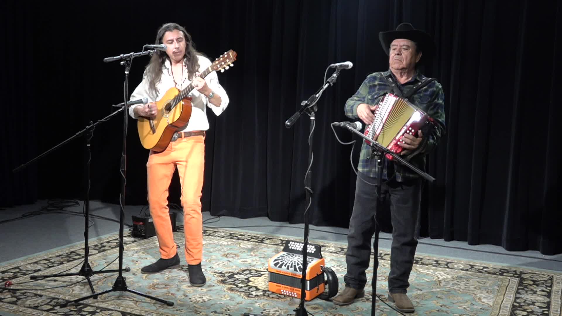 black background. Man standing in front of a mic to the right playing an accordian. Man in neutral colors standing in front of a mic playing a guitar.