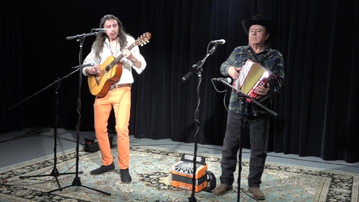 black background. Man standing in front of a mic to the right playing an accordian. Man in neutral colors standing in front of a mic playing a guitar.