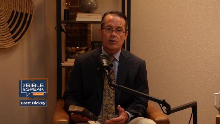 yellow toned room with a man in a suit seated in the middle in front of a podcast mic. Knick knack shelf in the back.