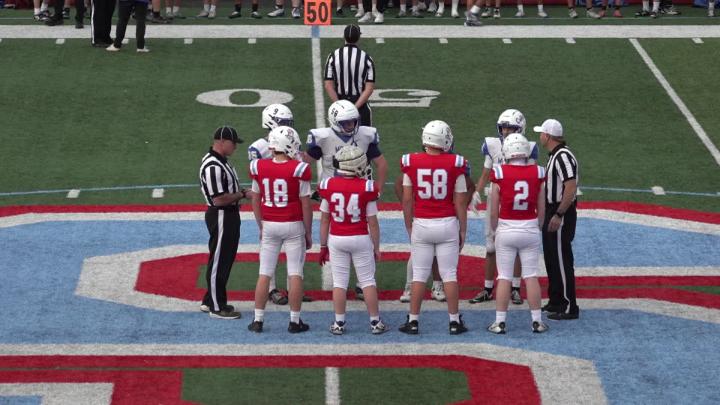 Football field with a large blue and red "S" in the center. Referees and players in red and white uniforms standing in the middle