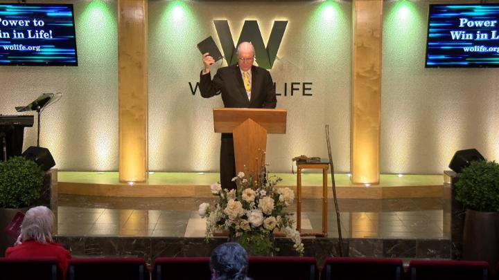 Man in a suit holding a bible next to a pulpit in the middle of a platform. Lit up white walls in the background with a large W and Word of Life in the center.