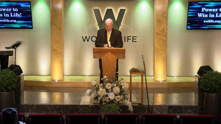 Man in a suit holding a bible next to a pulpit in the middle of a platform. Lit up white walls in the background with a large W and Word of Life in the center.