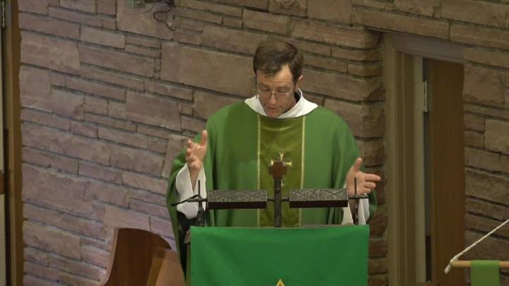 Priest dressed in green standing behind a pulpit. Brick wall in the back.