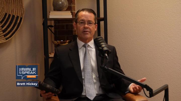 Yellow toned room. Man in a suit and glasses seated in the middle in front of a podcast mic. Knick knack shelf behind him.