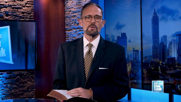 Man in a blue suit and glasses standing in the center holding a bible. Cityscape picture in the back right.