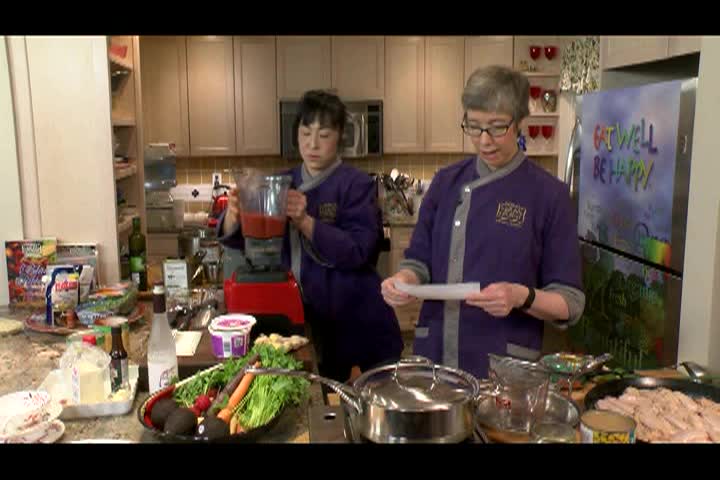 Woman on the right in a long purple chef jacket standing in front of a fridge and behind a kitchen counter. Woman in purple long chef jacket to left looking at various food items for a salad.