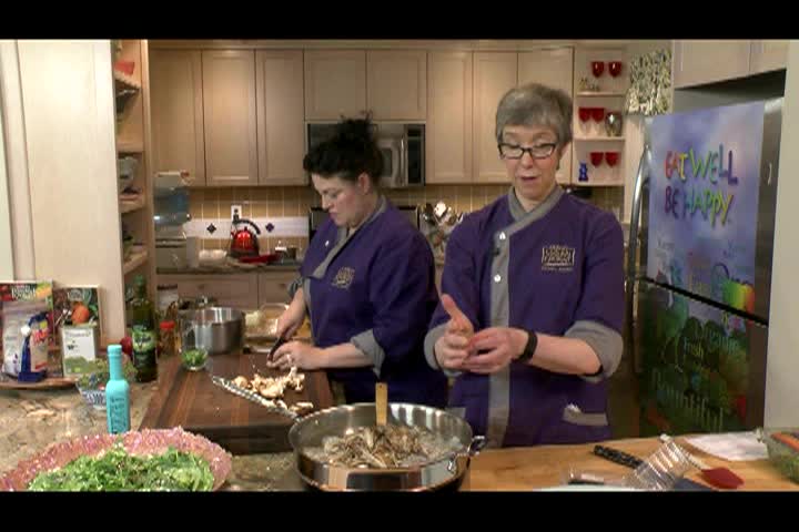 Kitchen setting. Woman with glasses and purple chef jacket to the middle right. Woman with dark hair and purple chef jacket to middle left preparing food.