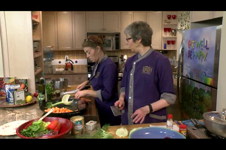 Kitchen setting. Woman with glasses and purple chef jacket to the middle right. Woman with dark hair and purple chef jacket to middle left preparing food.