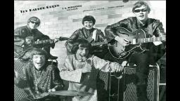 Black and white photo of a musical band. Three men in the background with guitars. One man in the front left missing a tooth and one man front center holding a microphone. The Barber Green on a brick wall in the background. 