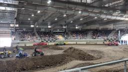 Photo inside an exhibit haul. People in stands in the background. Dirt hills with red, blue, and yellow demolition derby cars racing on dirt track.