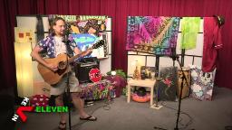 A man wearing a colorful shirt, shorts, and flip-flops plays an acoustic guitar and sings into a microphone in a decorated studio. Behind him are psychedelic posters, tapestries, and paintings with bright colors and patterns. To his right, a small table holds a lamp, and musical instruments including conga drums sit nearby. The bottom left corner of the screen shows the text “KEVIN ELEVEN.”